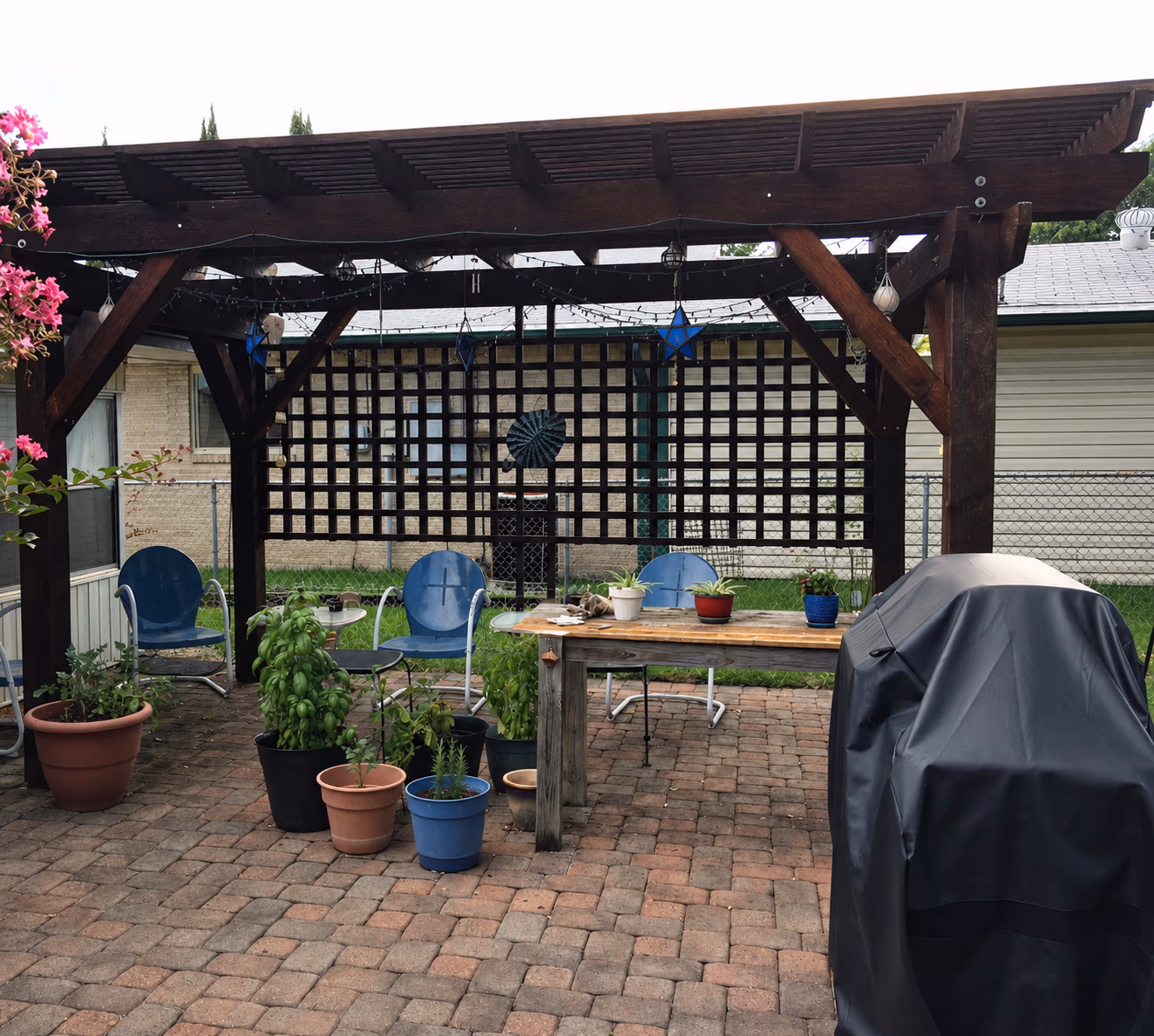 Backyard patio with wooden pergola, blue chairs, potted plants, a table, and a covered grill.