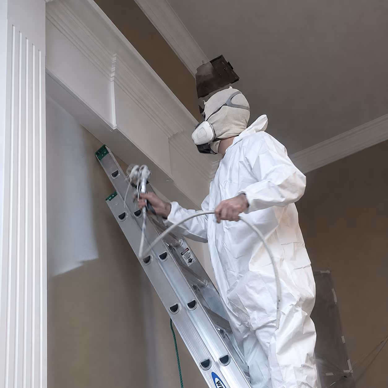 Person in protective suit and mask spray painting a white wall trim from a ladder indoors.