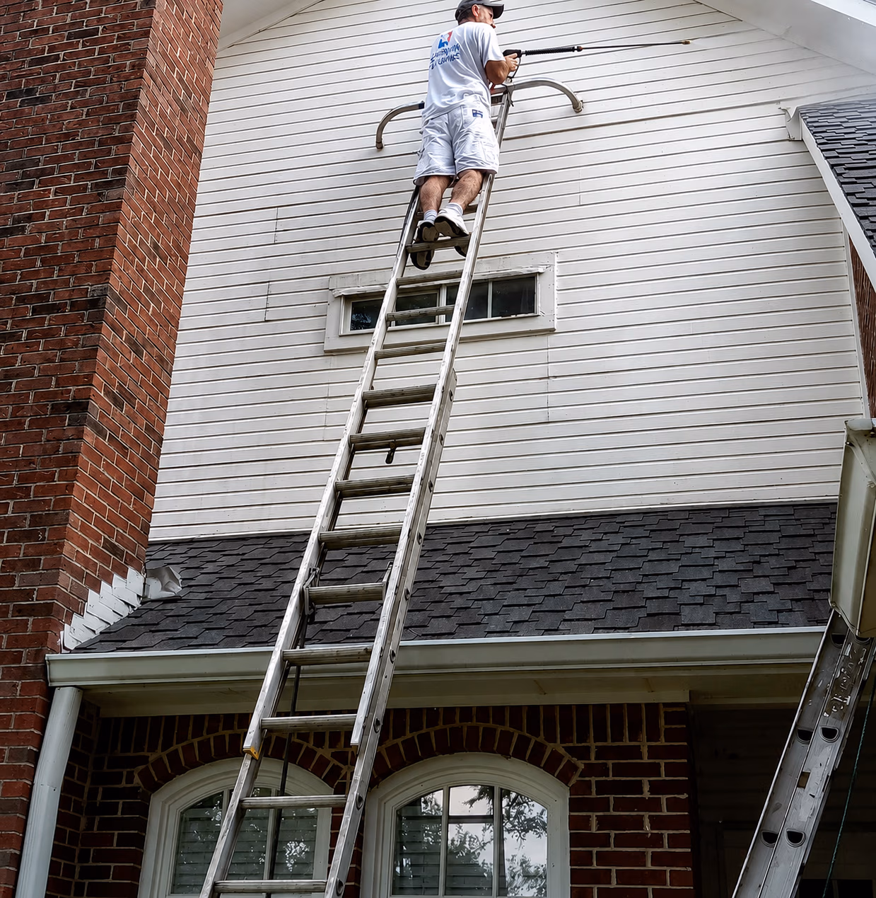 Man standing on ladder pressure washing white siding on the upper floor of a brick house.