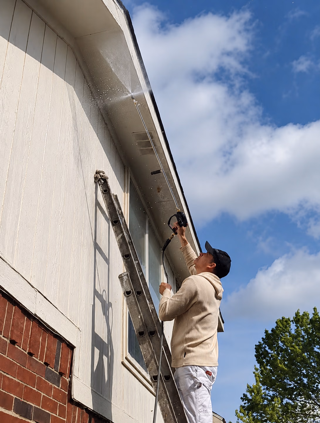 Man on ladder power washing the exterior of a white house under blue sky with clouds and trees.