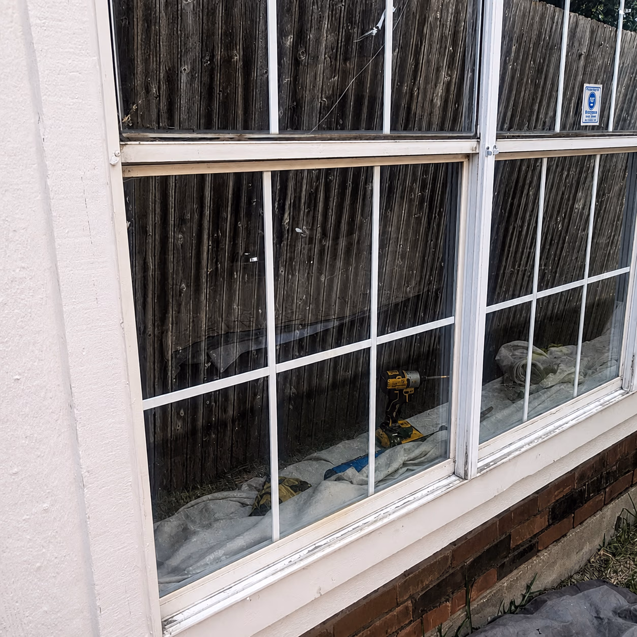 Window with white grid panes reflecting wooden fence outside; power drill and drop cloths inside.