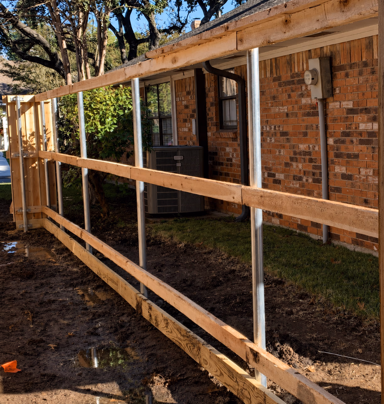 Unfinished wooden fence frame with metal posts beside a brick house with grass and trees.
