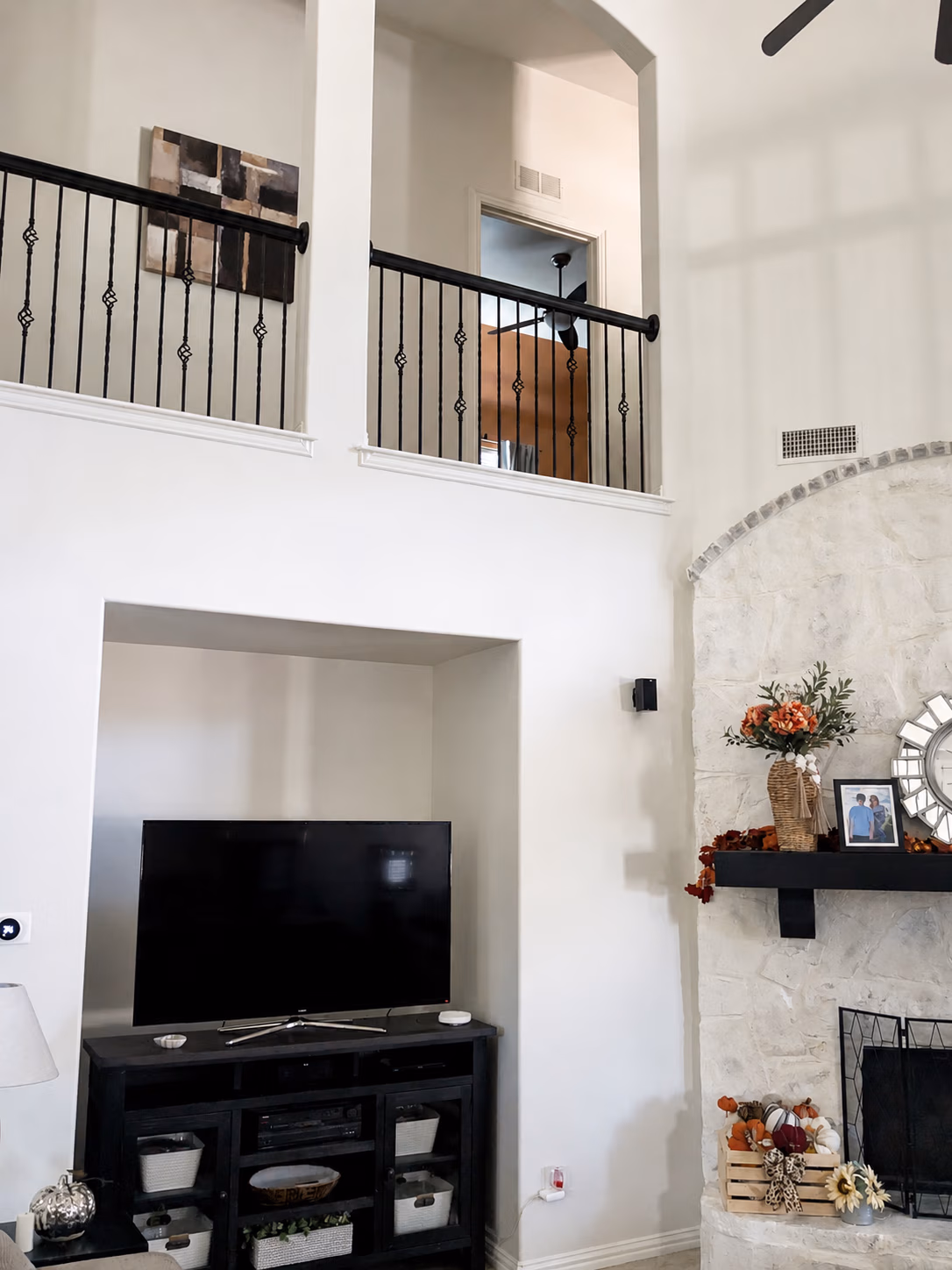 Living room with black TV on a cabinet, stone fireplace with fall decorations and a railing on second floor.