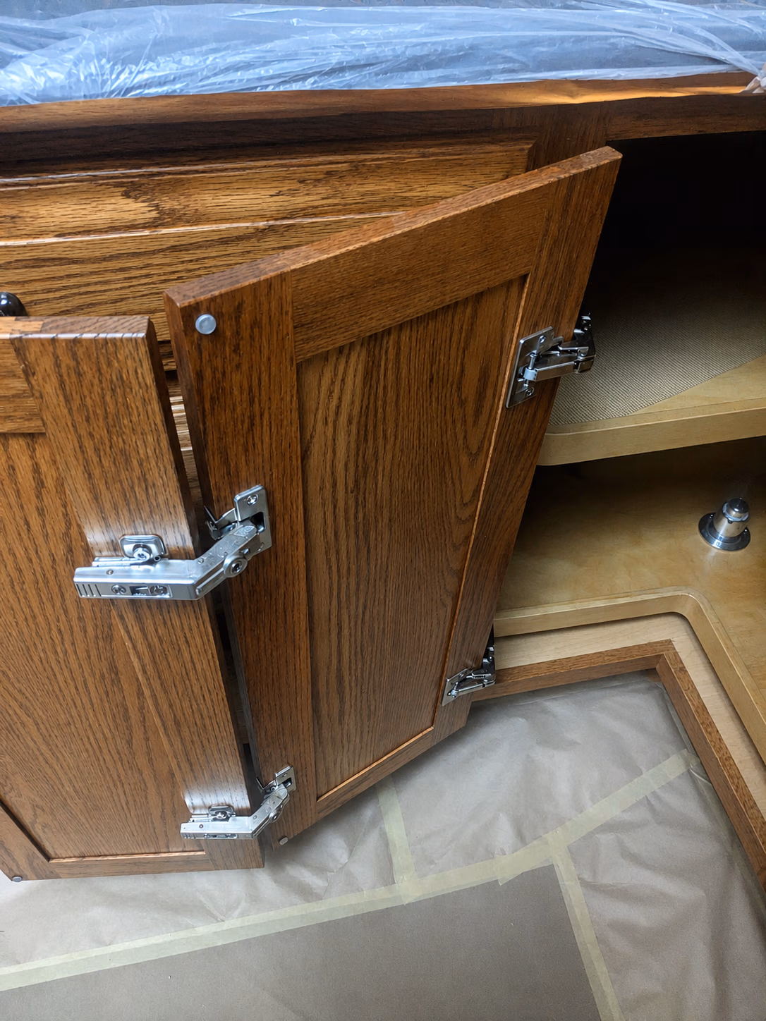Partially open wooden cabinet doors showing metal hinges inside a corner cabinet near taped floor.