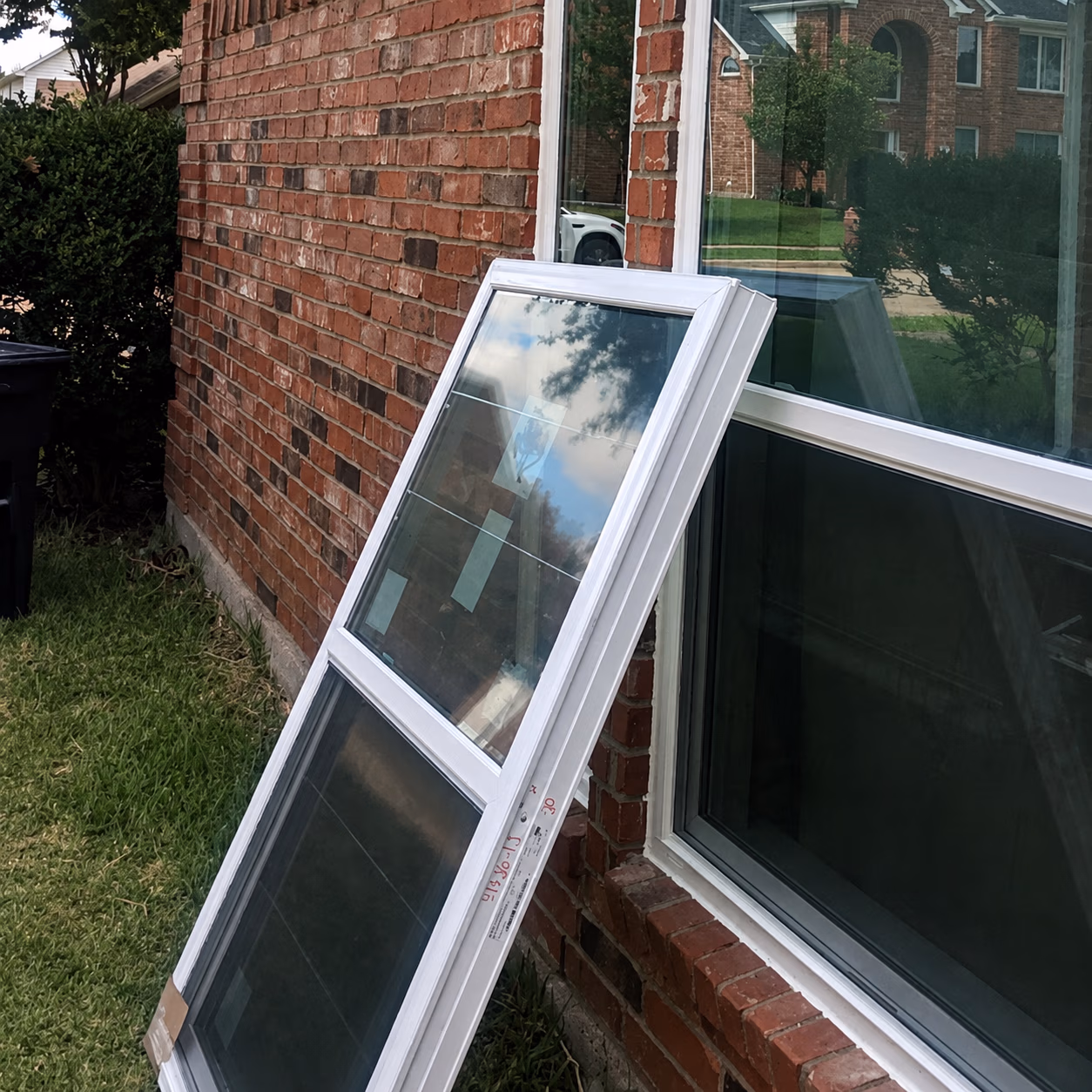 New white-framed window leaning against a brick house wall next to an existing window.