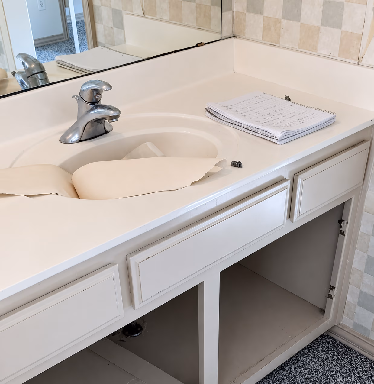 Bathroom sink with peeling vanity countertop and an open cabinet below, notebook on the counter.