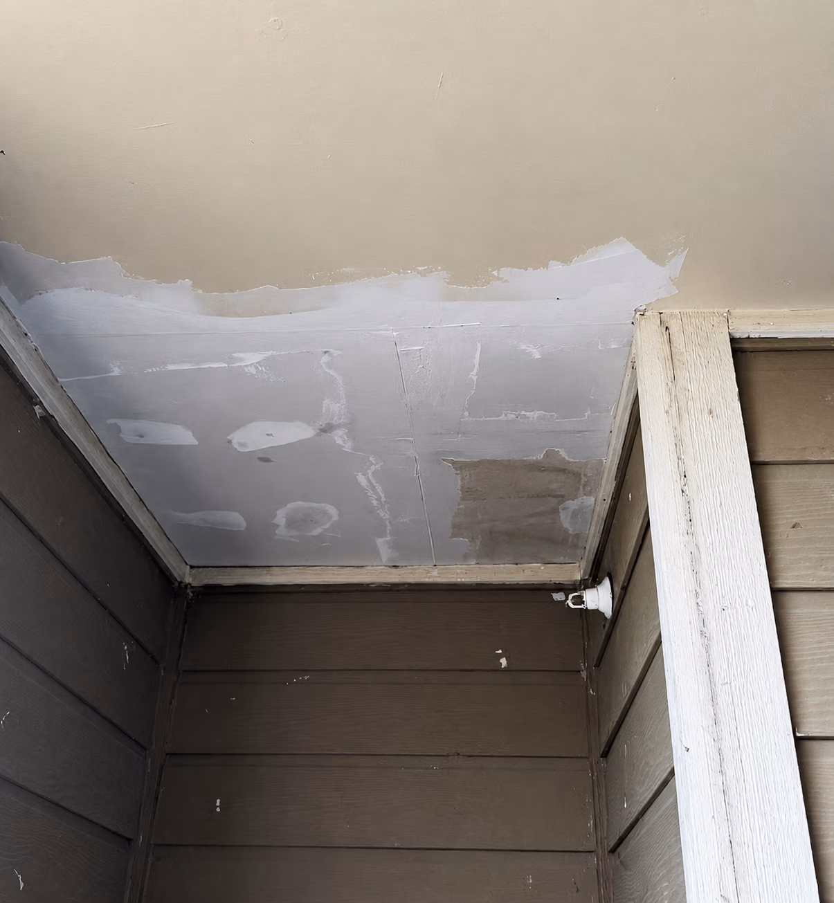 Ceiling with patches of white paint and spackle above brown paneled walls and a white corner beam.