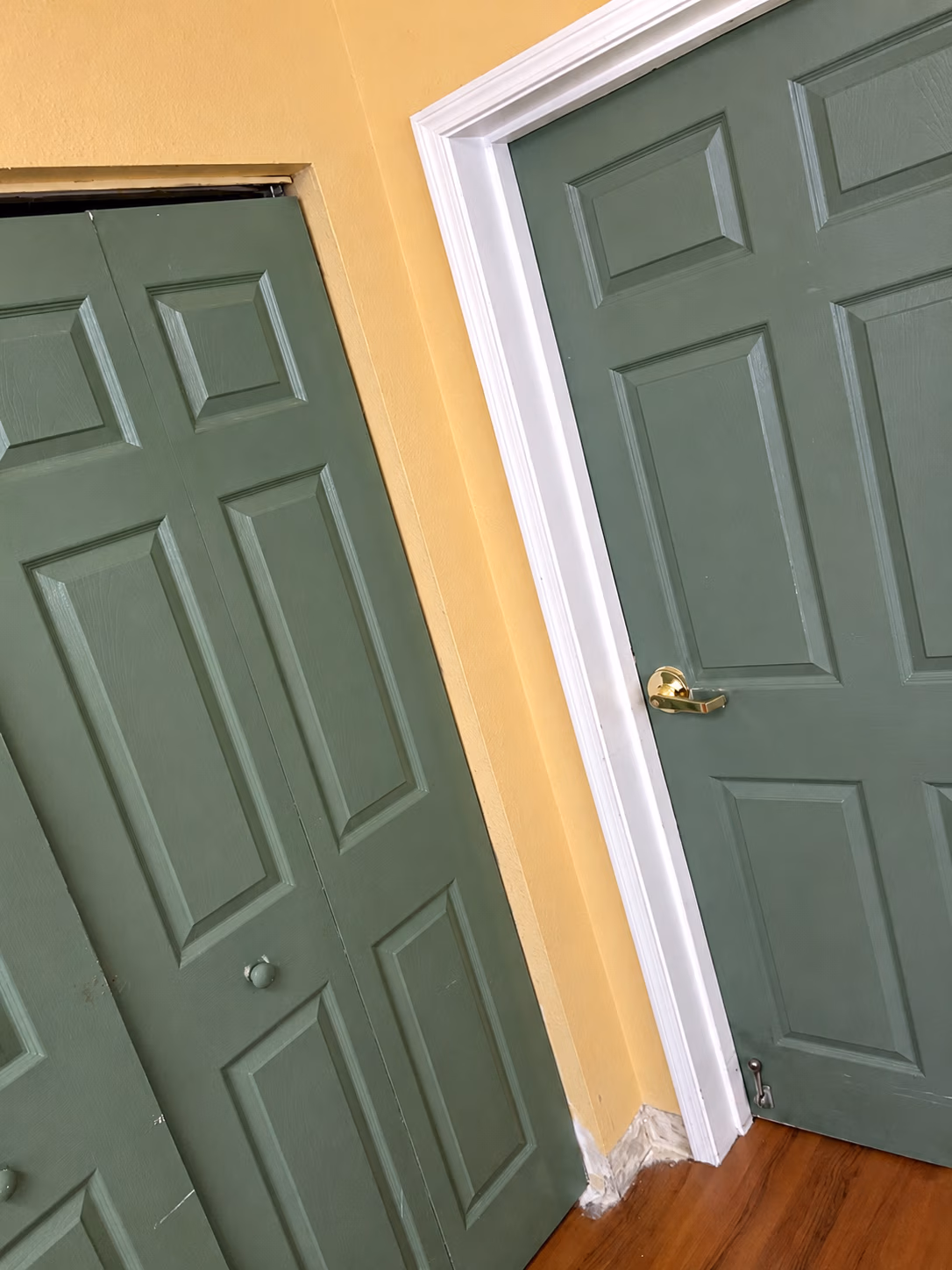 Two olive green paneled doors next to each other with yellow walls and wooden floor.
