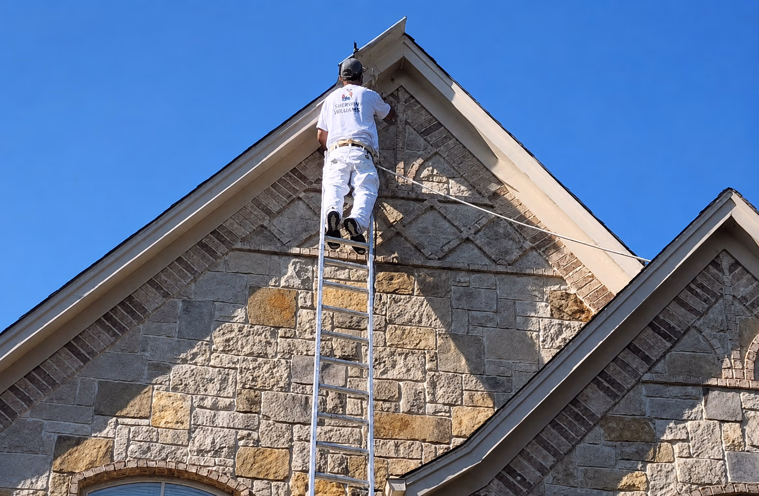 Painter in white uniform on ladder painting the stone exterior near house roof under clear blue sky.