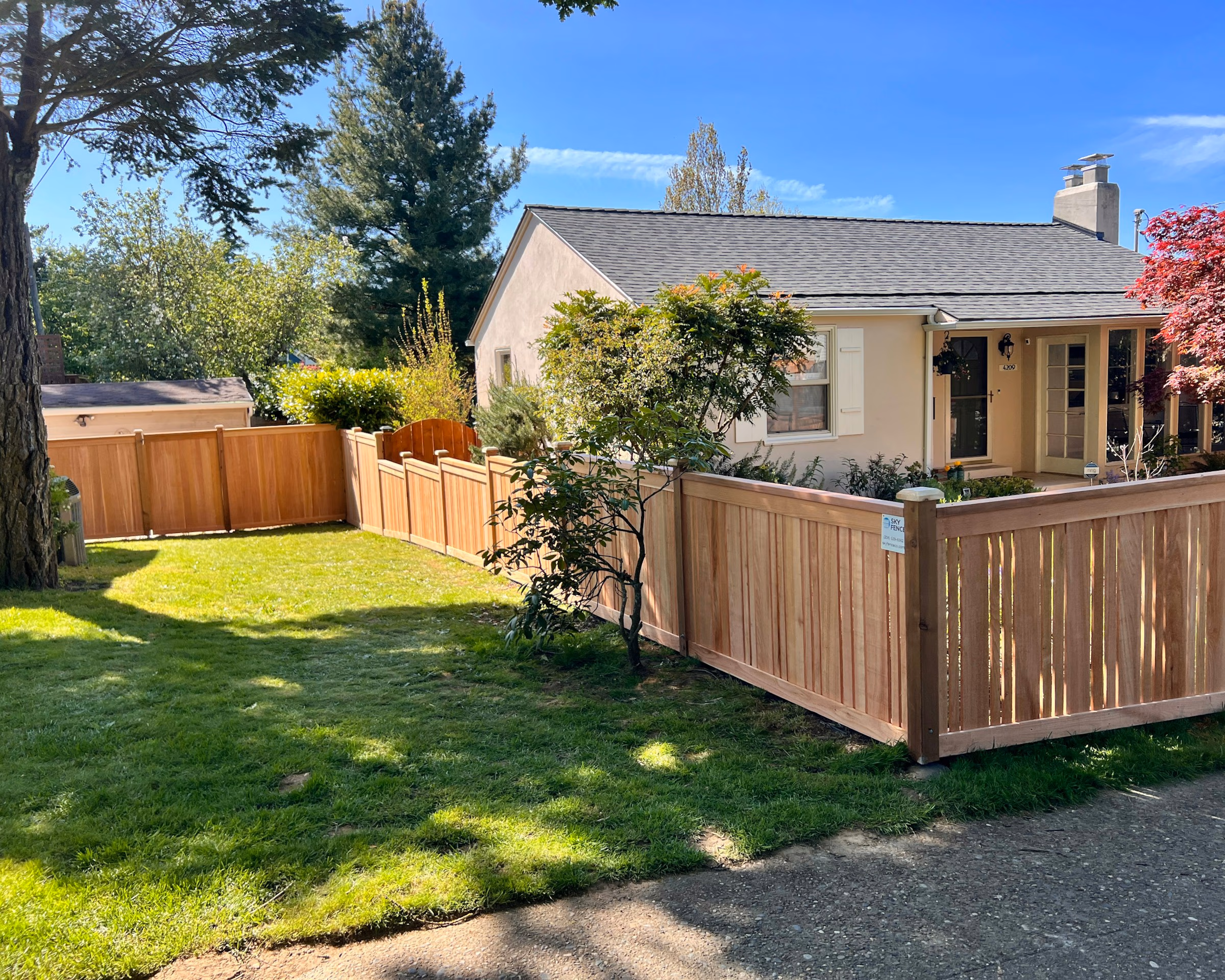 Small beige house behind a new wooden fence with green lawn and trees on a sunny day.