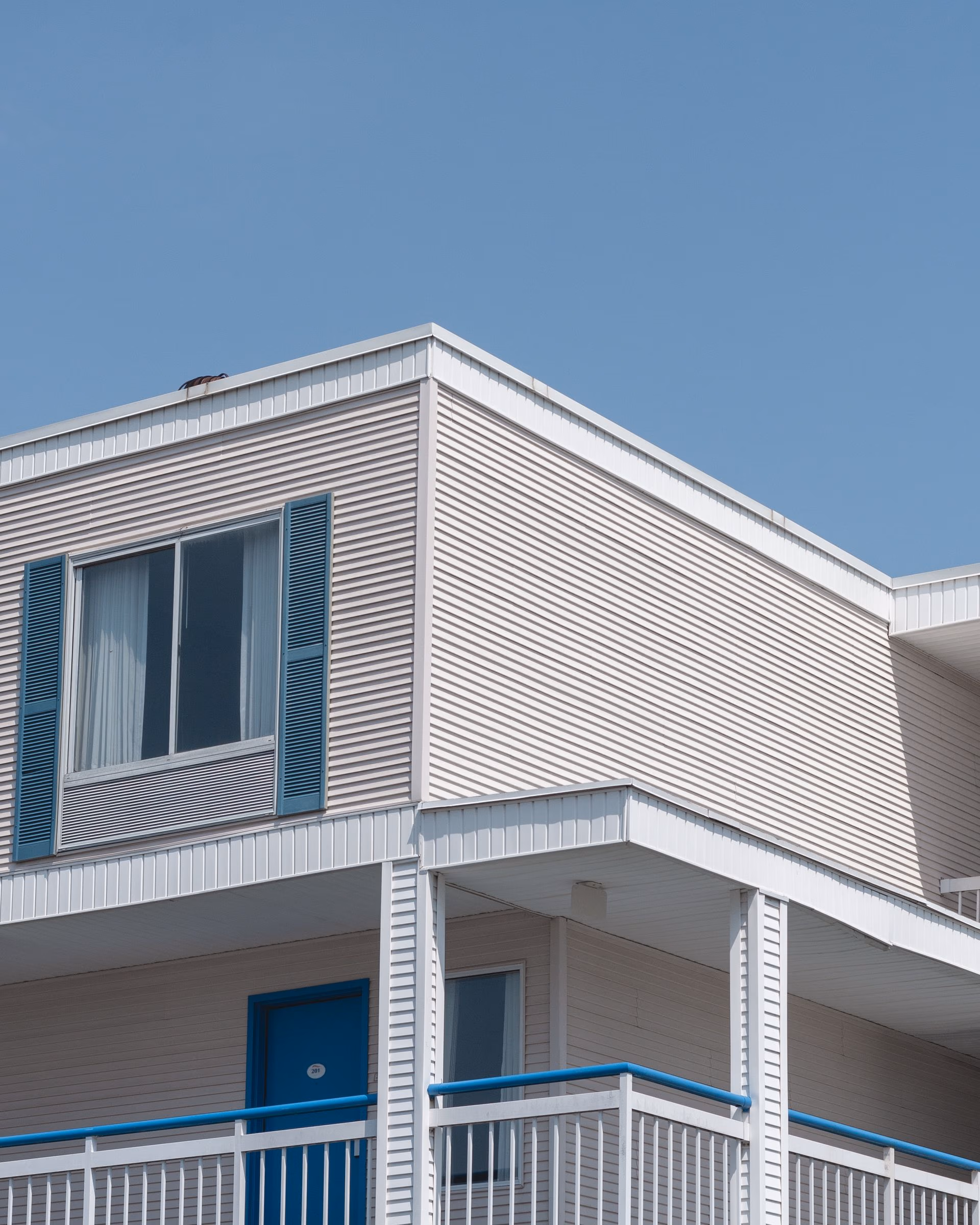 Exterior of a beige building with blue door, window shutters, and balcony railing under clear sky.