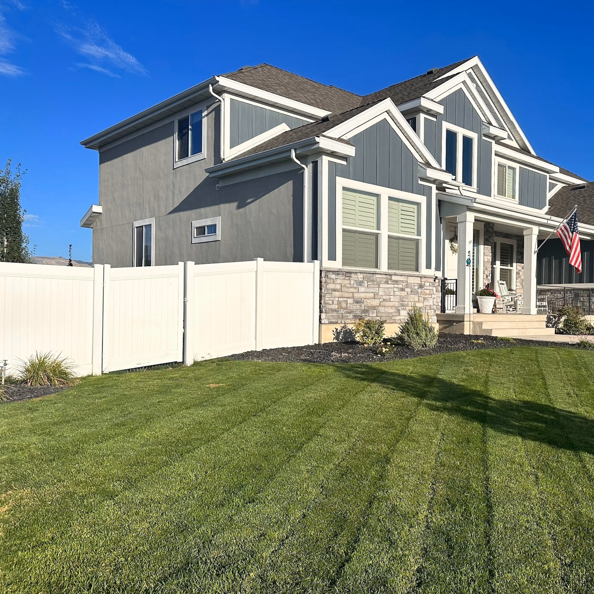 Two-story house with gray siding, stone accents, white fence, and a green lawn under a blue sky.